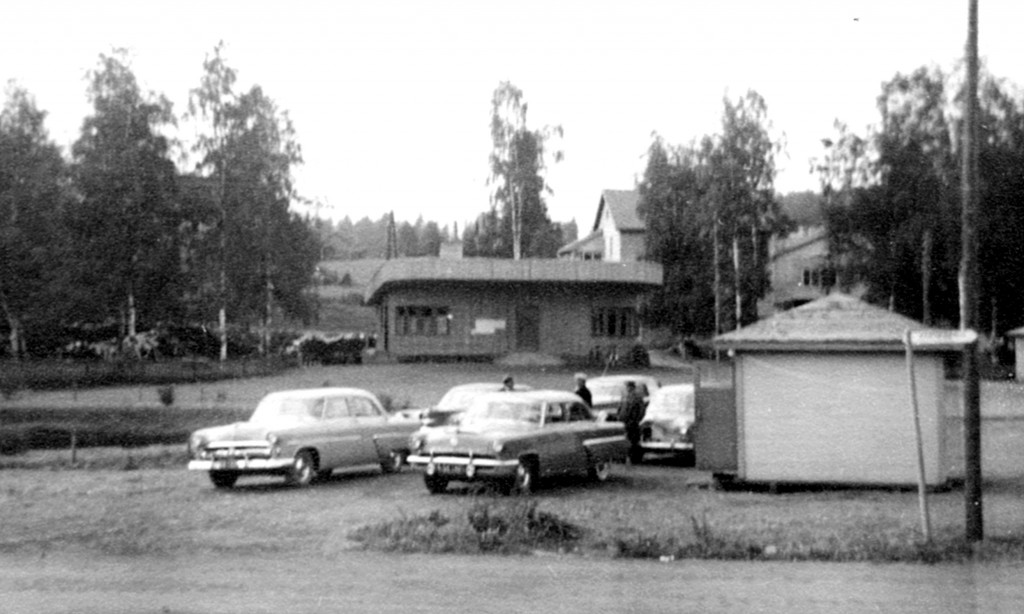1950-luvulla kirkonkylän taksit olivat enimmäkseen amerikanrautoja, mutta myös joitakin neuvostovalmisteisia esiintyi (Zil, Pobeda). Kuvan oikeassa reunassa näkyy osittin Matkahuollon koppi. Linja-autoaseman seutu oli vetistä aluetta, lähes suota. Alueen oikean reunan vieressä kulkenutta Häköojaa syventämällä saatiin maaperää kuivemmaksi, mutta täyttömaata oli tuotava runsaasti. Kuvan vasemmassa reunassa oleva lammikko on tehty kaivamalla. Sitä käytettiin kesäisin myös palokunnan vedenottopaikkana.